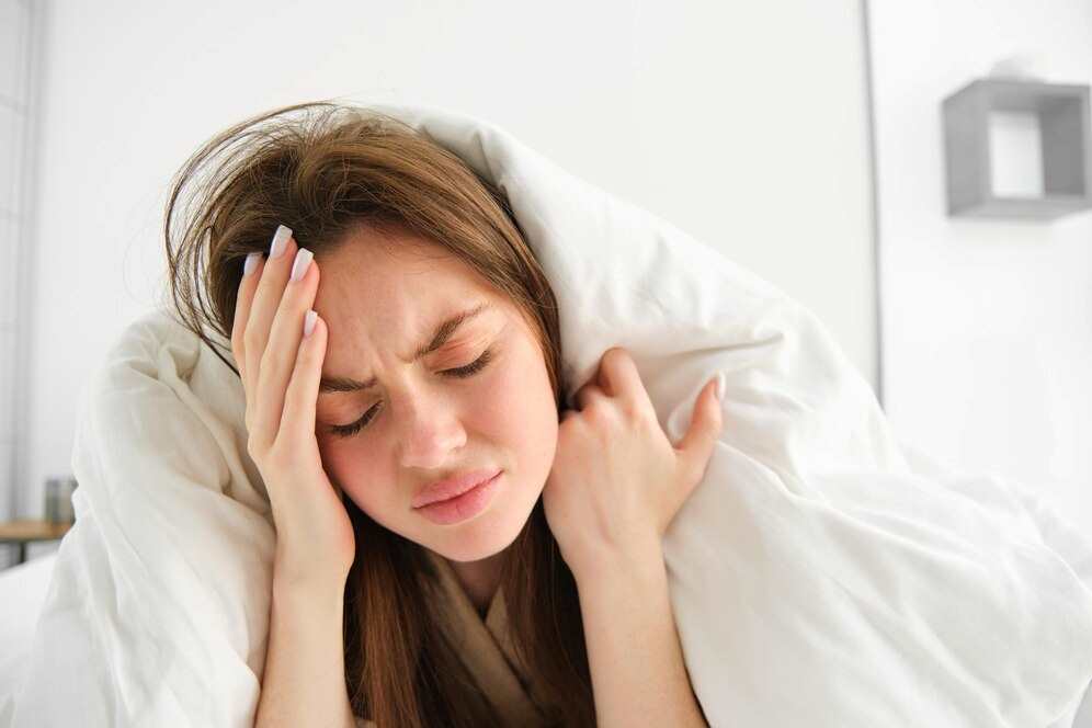 Žena, která má bolesti hlavy z chronické únavy, leží v posteli, přikrytá dekou